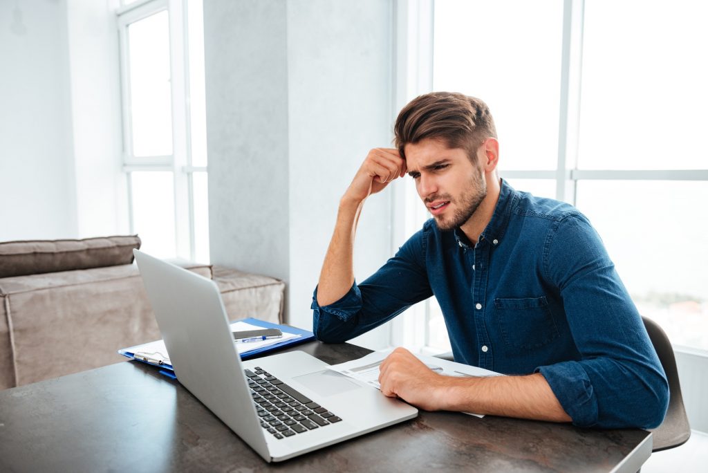 Sad young man sitting near laptop and holding head