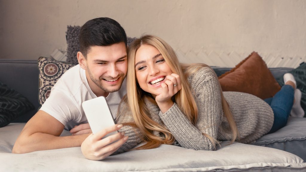 Loving young couple with mobile phone lying on sofa at home